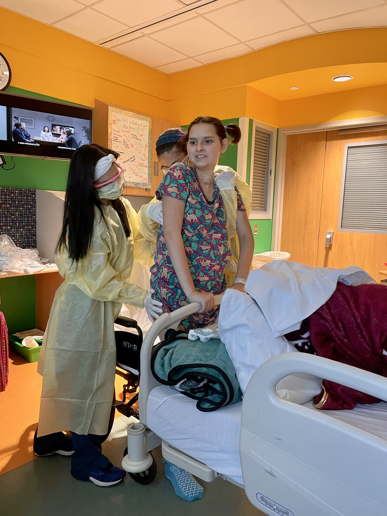Megan standing at the foot of her hospital bed for the very first time following the loss of her leg. There are two physical therapists helping to hold her.