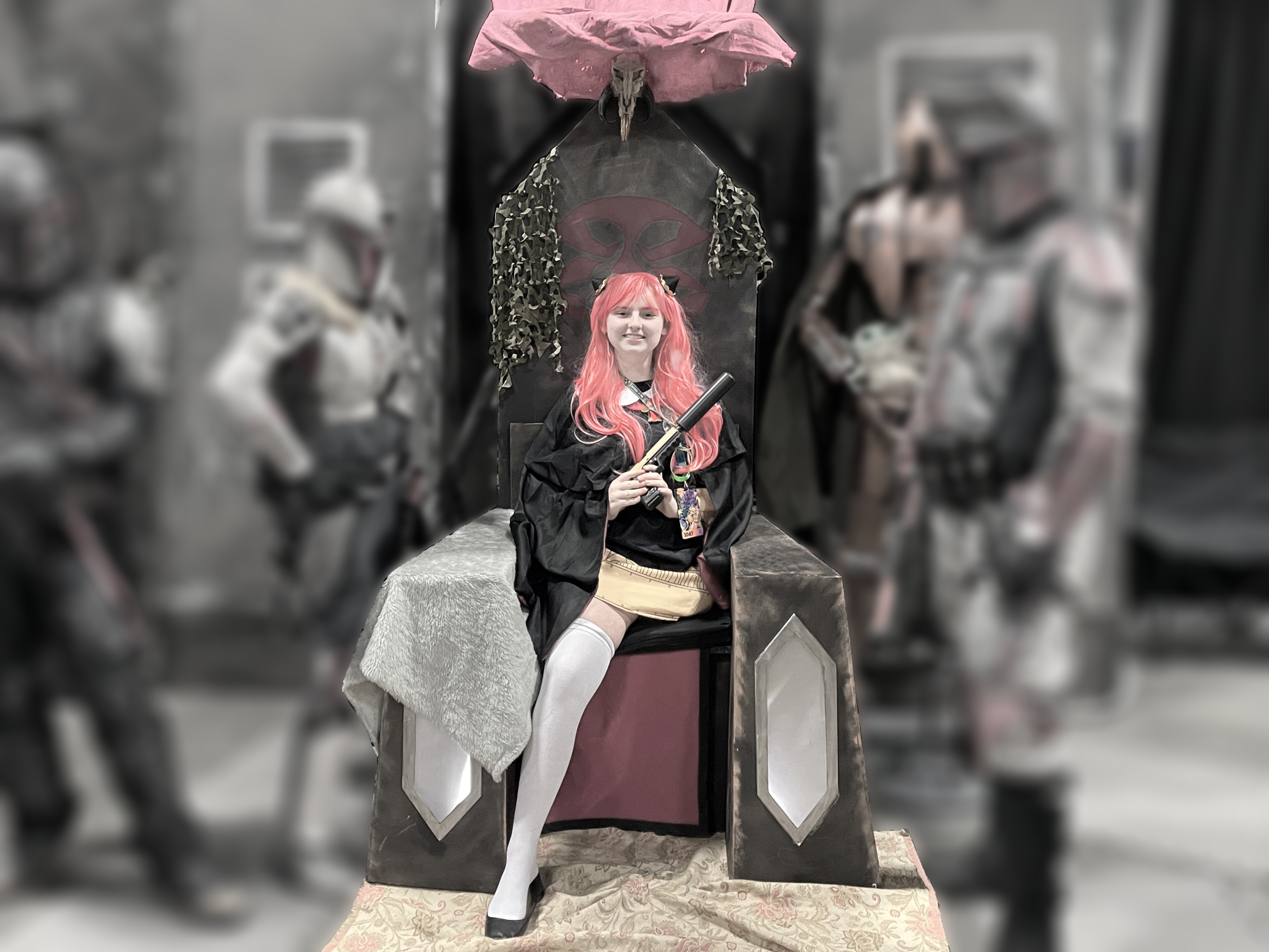 Megan in an cosplay as a teen age Anya from Spy X Family holding a pistol with a silencer while sitting on the Mandalorian throne.
